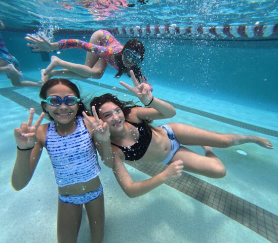 Three children underwater in a swimming pool, wearing swimsuits and goggles, make peace signs and smile. Bubbles surround them in the clear blue water.