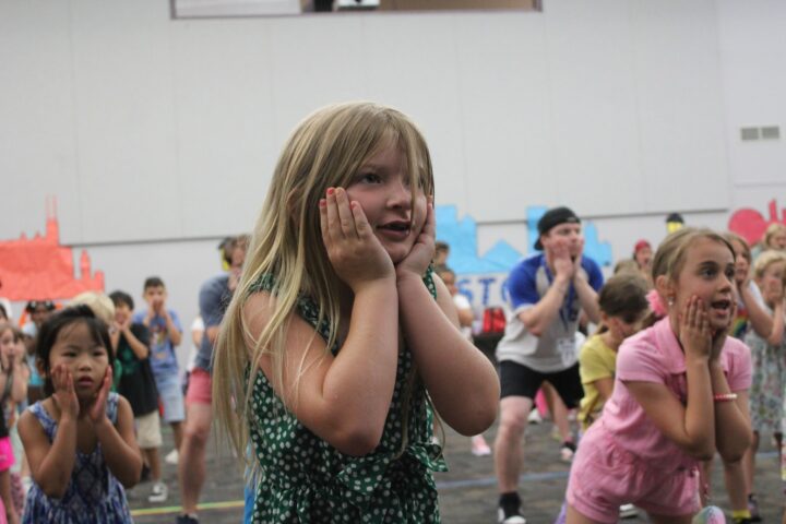 A group of children are engaged in a fun activity, with a young girl in the foreground holding her face with both hands.