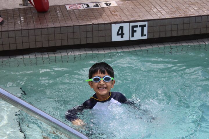 A smiling child wearing goggles and a swim shirt stands in a swimming pool marked 4 feet deep.