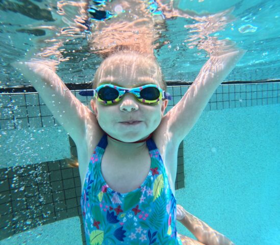 Child swimming underwater in a pool, wearing goggles and a colorful swimsuit, with arms stretched upwards.