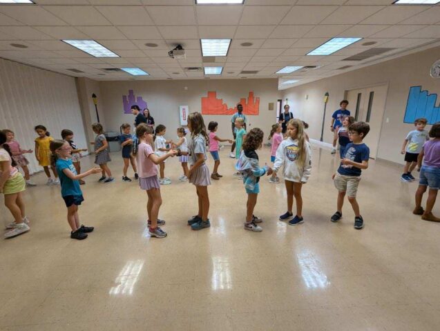 Children stand in pairs in a spacious room, engaging in activities. Several adults are present. Colorful wall decorations add to the lively atmosphere.