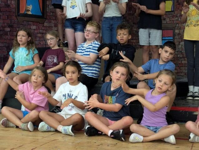 A group of children sitting on a stage, clapping their hands, with others standing behind them in a classroom setting.