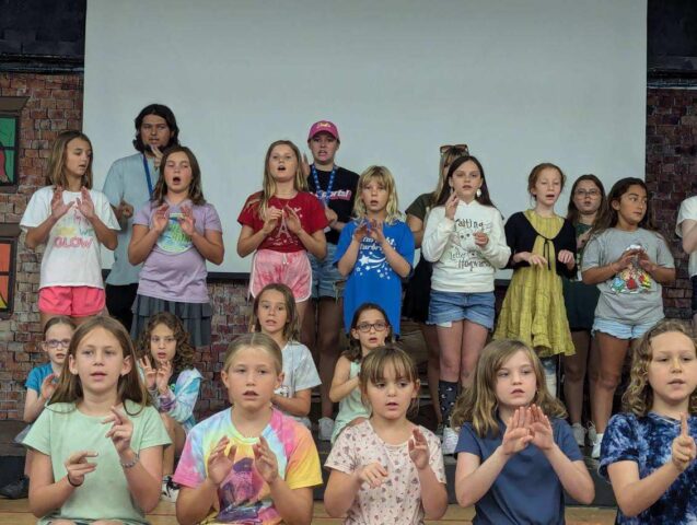 A group of children on stage performing a song with hand gestures, standing in rows against a plain backdrop.