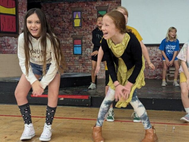 Children performing a playful dance on stage, smiling and energetically moving, with a brick wall backdrop and colorful window decorations.