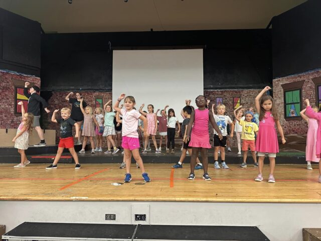 Children are on a stage practicing a group dance performance with arms raised. Some are wearing casual clothes, and the backdrop features colorful framed artwork.