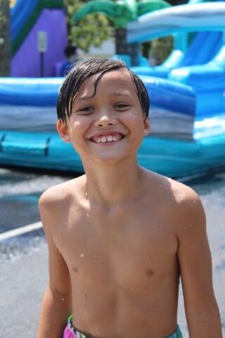 A smiling child with wet hair stands shirtless in front of a blue inflatable water slide.