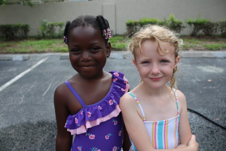 Two young girls in colorful swimsuits stand side by side on a paved area with greenery in the background.