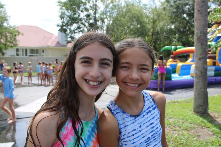 Two smiling girls in colorful swimsuits pose outdoors with a line of children and inflatable play structures in the background.