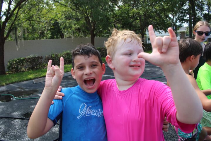 Two children smiling and making hand gestures, wearing wet shirts in a park. Trees and other kids are in the background.