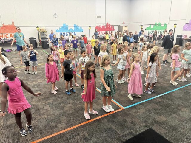 Children standing in rows indoors, participating in a group activity. Walls are decorated with city-themed banners.