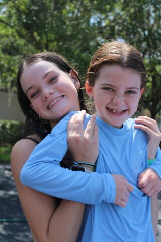 Two smiling people outdoors, one holding the other from behind. Both wear casual clothing. Trees are visible in the background.