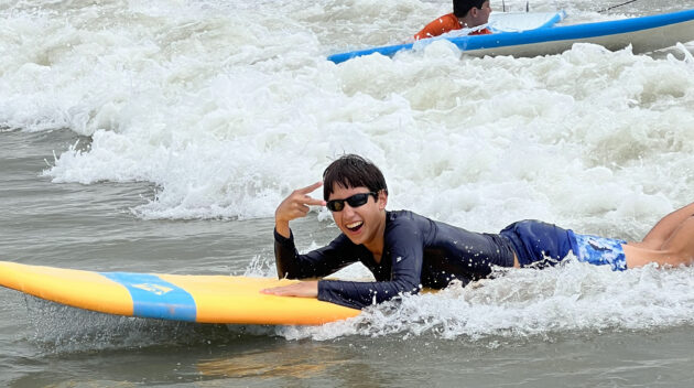 Boy laying on surfboard in the waves at the beach smiling during Camp Yalla
