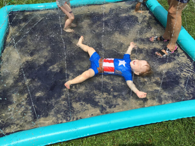 Child in patriotic swimsuit lies on wet inflatable water mat with spray, while another child's legs are visible nearby. Green grass surrounds the mat.