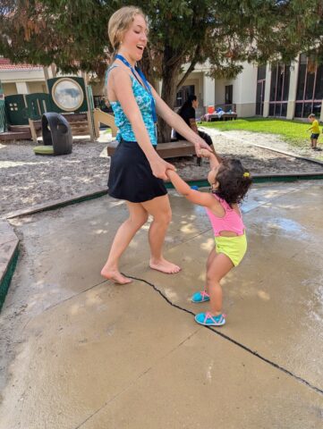 An adult and a toddler holding hands and playing outside on a paved area with a playground nearby.