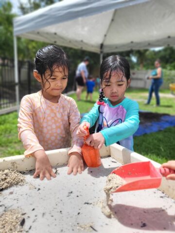 Two children play with sand and water in an outdoor area under a canopy. One holds a scoop, and the other looks at her hands. People are in the background.