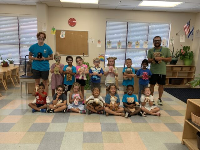 A group of children and two adults pose in a classroom, each holding a stuffed animal. The room features colorful flooring and bulletin boards.