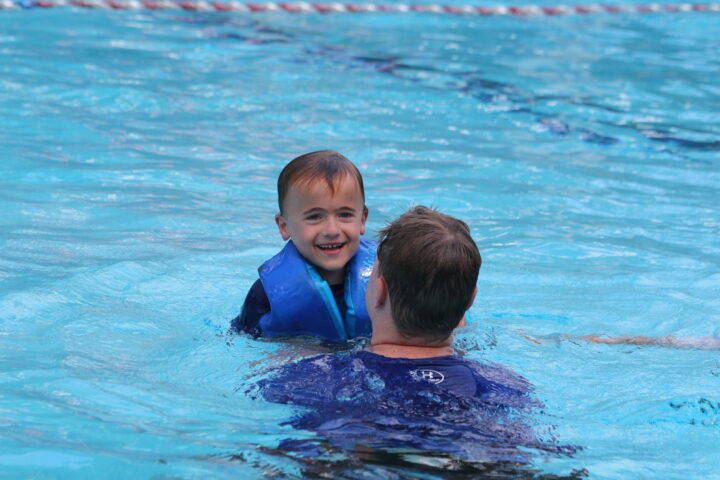 A child wearing a blue life vest smiles while being held by an adult in a swimming pool.