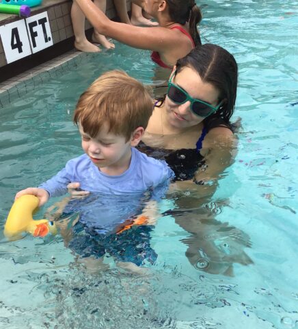 A woman in sunglasses helps a young boy play with a toy in a swimming pool.