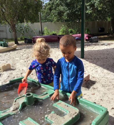 Two children play with water and sand at a green activity table in an outdoor playground surrounded by trees and sand.