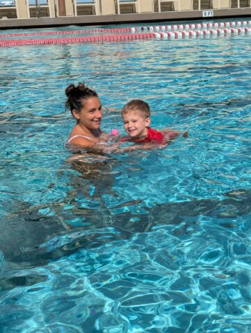 A woman and a child swim in a clear pool, with the child holding a pink ball.