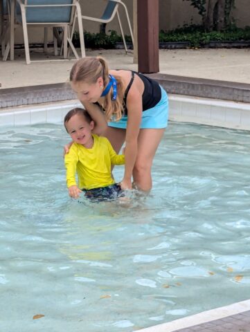 A woman helps a child in a yellow shirt stand in a swimming pool.