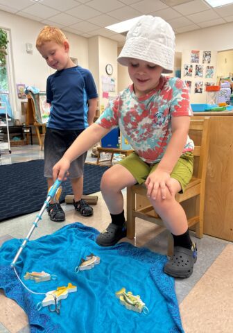 Two children in a classroom: one standing, one sitting with a fishing pole, pretending to fish toy sharks on a blue cloth.