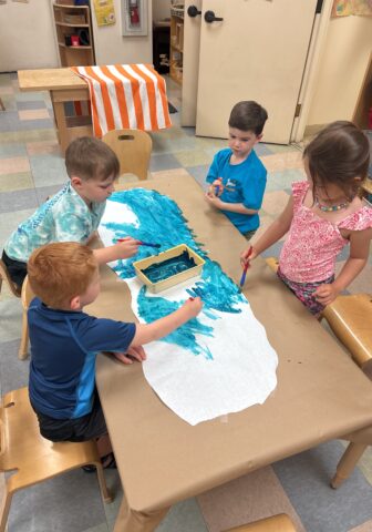 Children engaged in painting a large sheet of paper with blue paint at a classroom table.