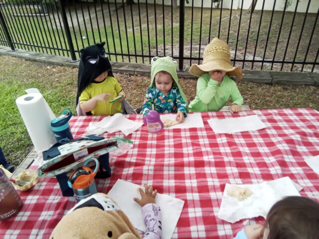 Children in costumes and hats sit at a red checkered tablecloth, eating snacks outdoors.