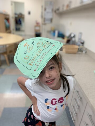 Young girl in a classroom wearing a paper hat with "Ranger Ned Fan" written on it, smiling at the camera.