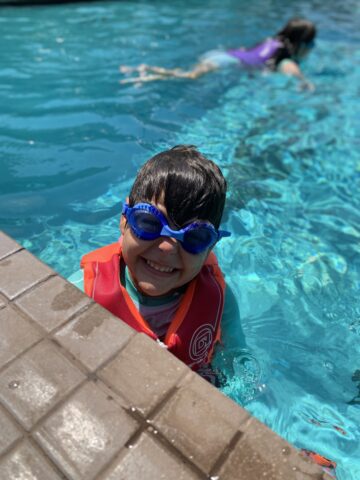 A child wearing a red life vest and blue goggles smiles at the pool's edge. Another person is swimming in the background.