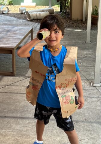 Child wearing a handmade cardboard vest, holding a cardboard tube to their eye like a telescope.