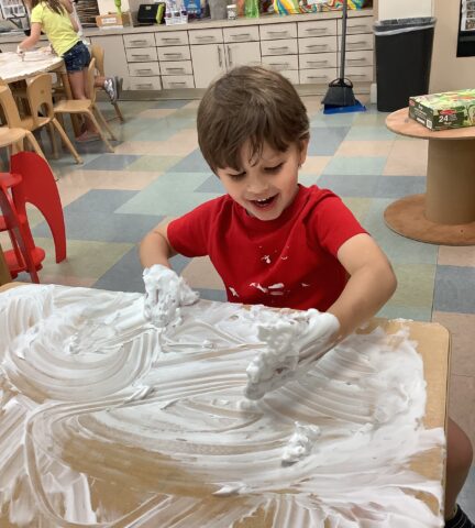 Child in a red shirt playing with foam on a table in a classroom.