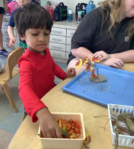 Child in a red shirt creates a clay craft with artificial leaves at a table, alongside a person working on a similar project. Paint supplies and other materials are visible.