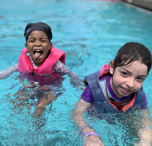 Two children wearing pink life jackets are in a swimming pool. One is playfully sticking out their tongue, and the other is smiling at the camera.