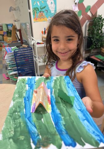 A child smiling and holding a painting with green, blue, and orange colors in a classroom setting.