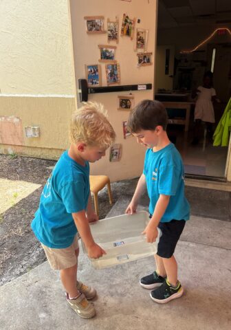 Two young boys in blue shirts carry a clear plastic container outside a building.