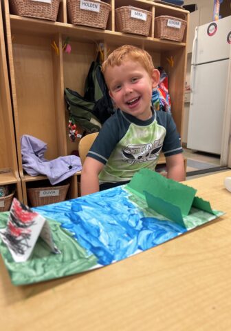 A child smiles while sitting at a table with a colorful craft project. Bins with names are visible in the background.