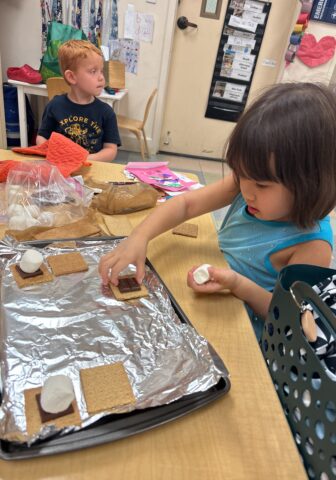 Two children make s'mores at a table. A girl places marshmallows on graham crackers and chocolate, while a boy sits in the background.