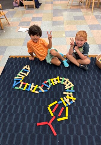 Two children sitting on a carpet showing peace signs, next to a design made from colorful wooden blocks.