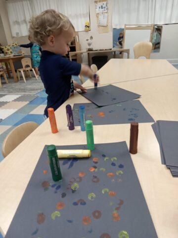 A toddler uses stamp markers on black paper at a classroom table, with several colored markers visible.