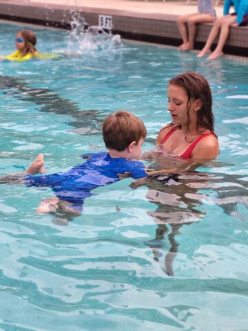 A young child in a blue swimsuit learns to swim with an instructor in a pool. Other children are swimming in the background.