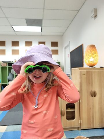 Child wearing a hat and holding green binoculars indoors, smiling and standing in a playroom with a lamp and cabinets in the background.