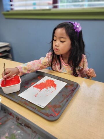 A young girl with a purple flower hair clip paints with red paint on paper using a stick, seated at a table.