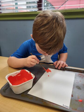 Child painting with red paint on white paper at a table.