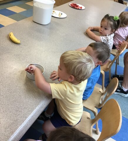 Children seated at a table, one child reaching into a bowl, with a banana and plates in the background.