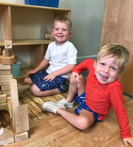 Two children are sitting on a wooden floor, smiling at the camera. They are surrounded by wooden blocks and a shelf in the background.