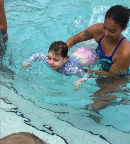 A child in a swim suit is being assisted by an adult woman in a pool. The child is wearing a long-sleeve rash guard with yellow fish designs.