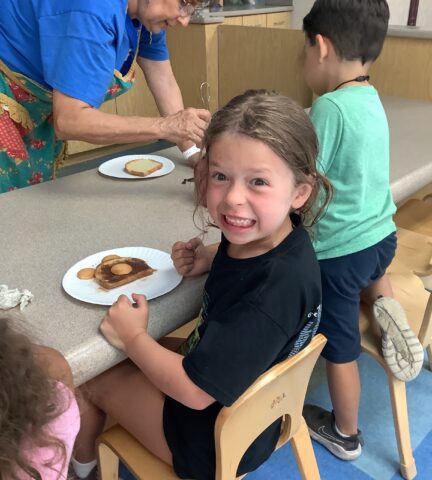 Child sitting at a table with a plate of pancakes, smiling broadly. Other children and an adult are in the background.