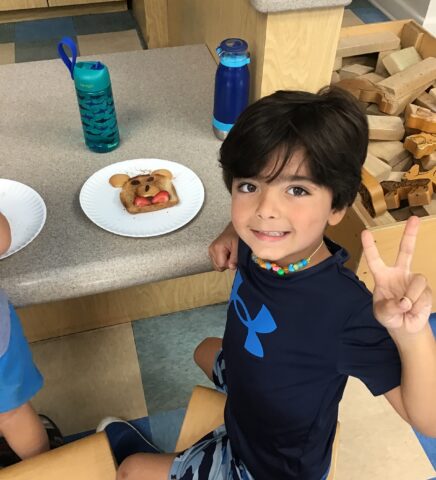 A child in a blue shirt sits at a table with a decorated piece of bread on a plate. He is smiling and making a peace sign with his fingers. Two water bottles are on the table.