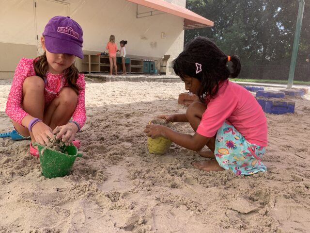 Two children wearing colorful clothing play with sand using buckets at an outdoor playground. Other children are visible in the background near a building.